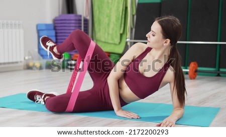 a woman in a burgundy tracksuit does leg exercises with gymnastic elastic bands on the side. video and online lessons for sports. clothes and shoes for gymnastics. professional fitness trainer.