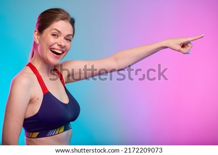 Happy emotional sporty woman with mouth open dressed fitness sportswear pointing finger to side empty space. Female fitness portrait isolated on neon multicolor background.