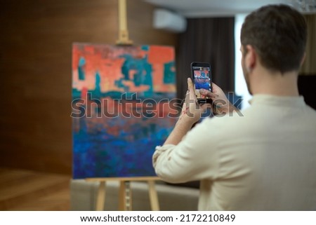 Man artist takes pictures by phone masterpiece of modern art, contemporary abstract painting for social media, rear view Royalty-Free Stock Photo #2172210849