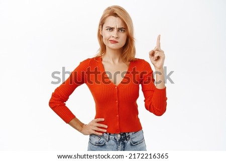 Woman points up with suspicious face expression, has doubt or disbelief, stands over white background in casual outfit