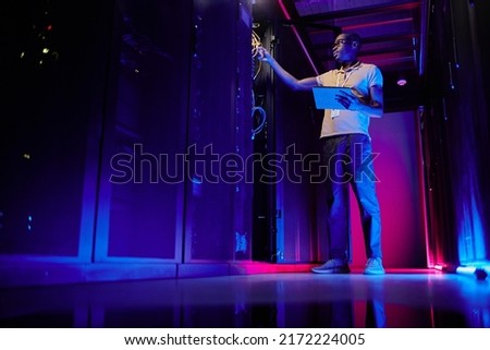 Full length portrait of African American system administrator setting up server network in data center lit by neon light, copy space Royalty-Free Stock Photo #2172224005