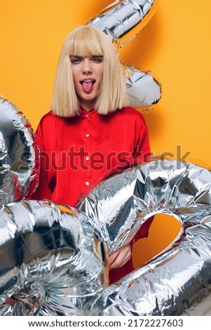 a bright vertical photo on a yellow background of a happy woman with balloons in the form of silver numbers, obliviously showing her tongue to the camera
