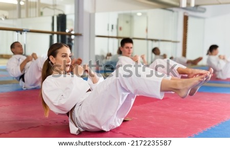 Sporty young woman in white kimono warming up before group martial arts training in gym, doing abdominal crunch.. Royalty-Free Stock Photo #2172235587