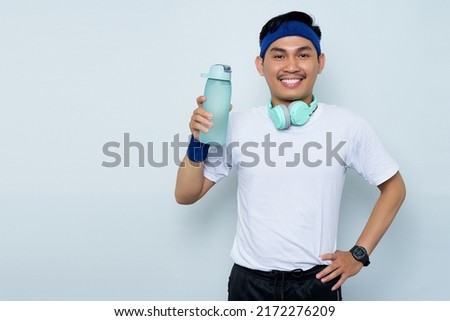 Portrait of Smiling young Asian sporty man in blue headband and white t-shirt with headphones, Holding water bottle isolated on white background. Workout sport concept