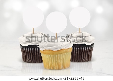 Three classic white frosted cupcakes chilling atop an elegant marble tabletop surrounded by glowing white lights. Royalty-Free Stock Photo #2172283743