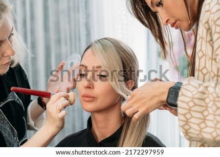 A hairdresser and a makeup artist together prepare a beautiful model for a photo shoot. The girl gets her hair and makeup done in a beauty salon.