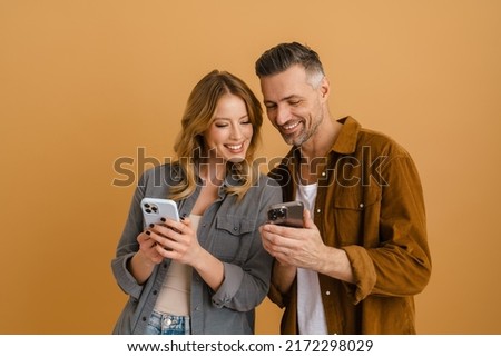 White happy couple using mobile phones while laughing isolated over beige background