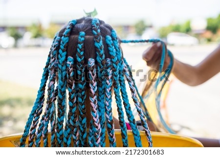A little waiting tanned girl in a bright yellow summer suit, weaves colored pink-blue African braids in her dark wet thick hair on a hot sunny sea day Royalty-Free Stock Photo #2172301683