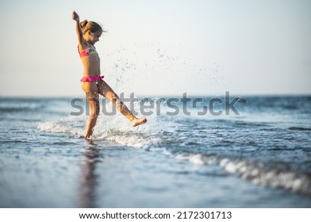 Funny slender cheerful girl playing with small waves, kicking their feet and spinning in place under the warm bright summer sun enjoying the long-awaited vacation