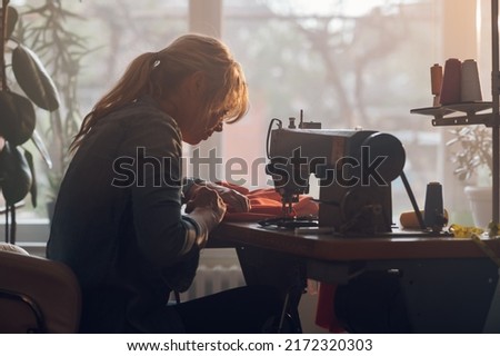 Silhouette of a beautiful middle aged tailor woman sewing on a sewing machine while sitting at her moody working place at home. Fashion atelier, tailoring, handmade clothes concept. Slow Fashion.