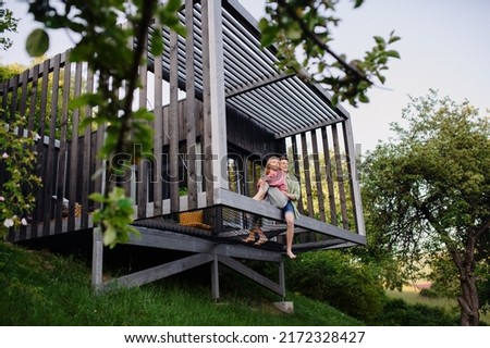 Young couple sitting and cuddling in hammock terrace in their new home in tiny house in woods, sustainable living concept. Royalty-Free Stock Photo #2172328427