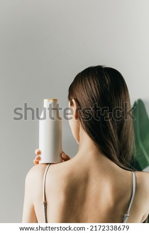 Woman holds a white bottle of cosmetics in her hand, standing with her back to the camera. Mock up shampoo, gel. Royalty-Free Stock Photo #2172338679