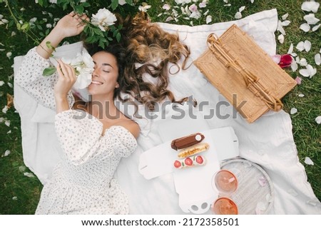 Young beautiful woman with long curly hair wearing white dress having picnic with champagne and fresh eclairs in blooming rose garden.