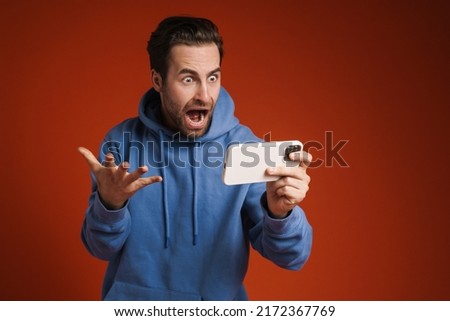 Young shocked man screaming while playing online game on cellphone isolated over red background