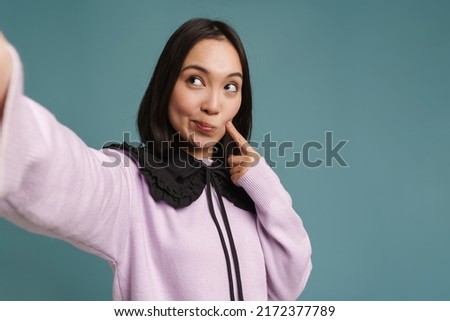 Asian woman pointing finger at her cheek while taking selfie photo isolated over blue background