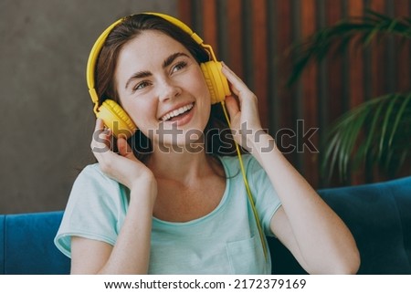 Young dreamful pensive woman she 20s wear casual clothes mint t-shirt hold use mobile cell phone headphones listen to music in reverie mood sit on blue sofa indoor rest at home in own room apartment.