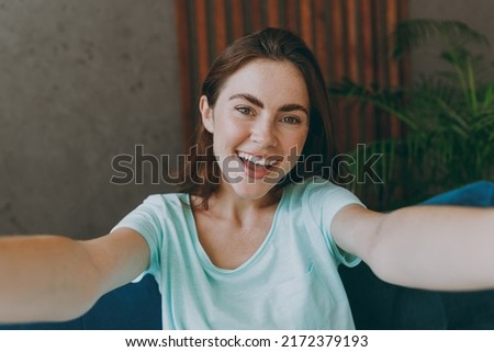 Close up young happy excited woman she 20s in casual clothes mint t-shirt doing selfie shot pov on mobile cell phone sit on blue sofa indoor rest at home in own room apartment People lifestyle concept