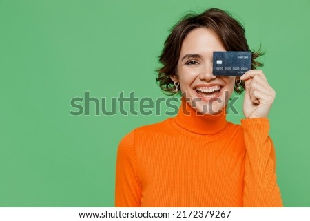 Young smiling happy cheerful fun woman 20s wear orange turtleneck hold in hand cover eye with credit bank card isolated on plain pastel light green background studio portrait. People lifestyle concept