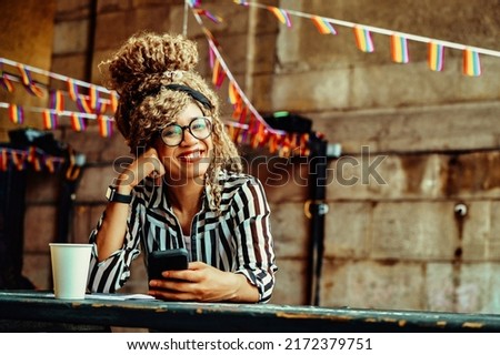 Portrait of smiling woman with afro hairstyle, eyeglasses, smartphone and paper cup looking at camera sitting in a coffeeshop shot