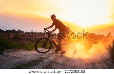 Cyclist on a gravel bike rides along the trail raising dust from the rear wheel during framatic sunset.