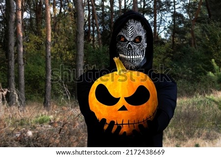 Grim reaper holding halloween pumpkin head. Man in death mask with fire flame in eyes on dark autumn forest pine tree background. Halloween holiday concept. Dark horror.