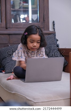 A little girl sitting on the sofa doing her homework on the laptop; serious. 