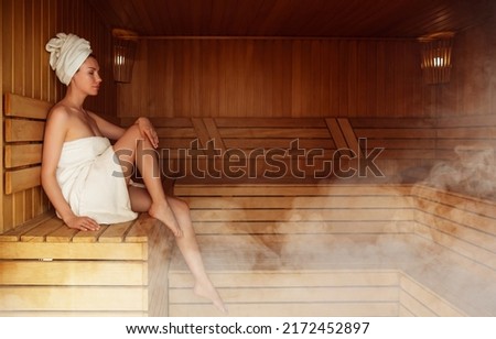 Young woman relaxing and sweating in hot sauna wrapped in towel. Girl In Sauna. Interior of Finnish sauna, classic wooden sauna with hot steam. Russian bathroom. Relax in hot bathhouse with steam. Royalty-Free Stock Photo #2172452897