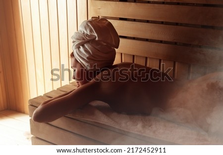 Young woman relaxing and sweating in hot sauna wrapped in towel. Girl In Sauna. Interior of Finnish sauna, classic wooden sauna with hot steam. Russian bathroom. Relax in hot bathhouse with steam. Royalty-Free Stock Photo #2172452911