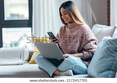 Shot of beautiful woman working with her laptop while using mobile phone sitting on a couch at home. Royalty-Free Stock Photo #2172457705