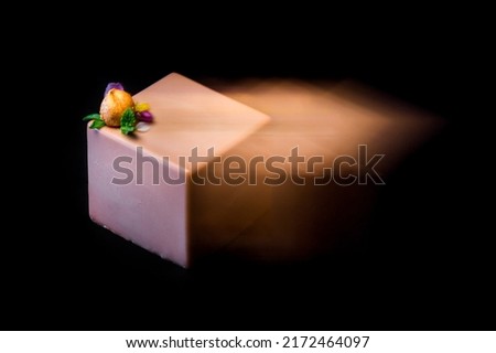 On a black background lies a square piece of cake in frosting with hazelnuts, a flower and a mint. Long exposure blurred photo