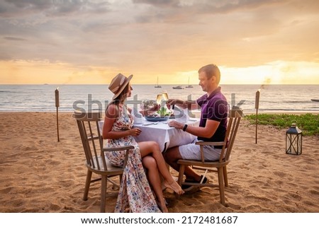 Couple in love drinking champagne wine on romantic dinner at sunset on the beach with yachts on background. Young couple celebration anniversary, clink glasses at served table on sea sandy coastline. Royalty-Free Stock Photo #2172481687