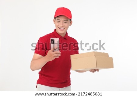 Portrait of handsome young delivery man checking order by smart phone, holding cardboard box to deliver to customer, in red shirt uniform, isolated on white background