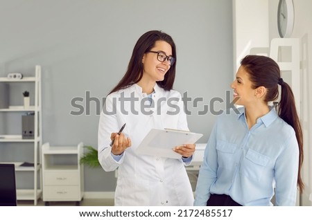 Friendly cheerful nurse or doctor at modern clinic talking to patient. Happy physician or gynaecologist holding clipboard, smiling and giving medical consultation to young woman. Health concept Royalty-Free Stock Photo #2172485517