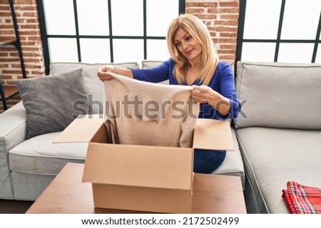Middle age blonde woman sitting on the sofa opening a box with sweater relaxed with serious expression on face. simple and natural looking at the camera. 