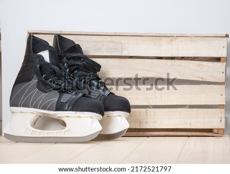 A pair of black hockey skates with well-sharpened metal blades in a white plastic frame designed to play ice hockey as a popular winter sport stay next to the craft wooden wall. Turned to the right.