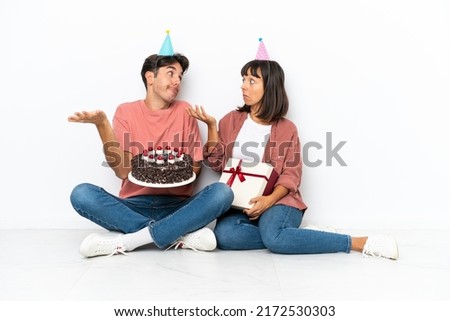 Young mixed race couple celebrating a birthday sitting on the floor isolated on white background making unimportant gesture while lifting the shoulders