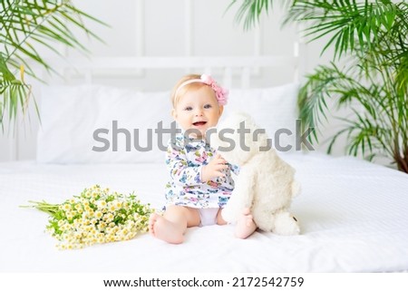 A cute little baby in a dress made of natural cotton fabric is sitting on white bed linen in the bedroom. A little happy child in bed with a teddy bear and a bouquet of daisies