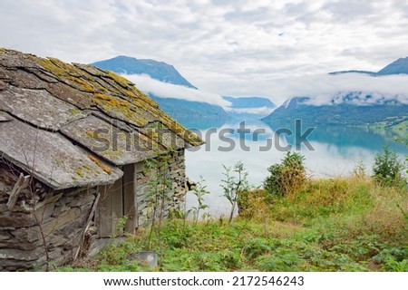 Rural landscape in Norway. Sea shore, old boathouse, covered with mossy stone tiles, old road, white cloud above the water in the Lustrafjord Royalty-Free Stock Photo #2172546243