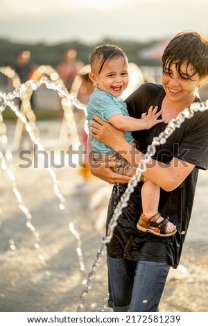 children run around the fountain. The older brother is holding the younger one in his arms. Children have fun in the park on a hot summer day