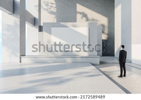 Businessman in suit back view looking at blank white banner with place for your logo or text on light grey wall in sunlit spacious hall with concrete design, mock up