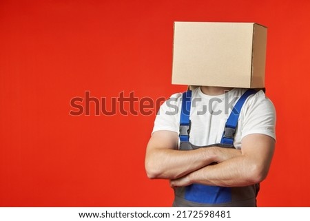 Anonymous loader confidently stands in a work suit with straps on a red background with a box on his head. A professional male loader thinks only about the safety and security of the transported items