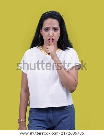 Beautiful Indian girl or Young south Asian woman saying shh or keeping a secret with angry expression.asking for silence and quiet, gesturing with finger in front of mouth. Over yellow background.