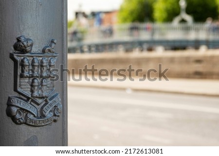 Coat of arms symbol of Dublin on a metal lamp post in focus. Ha'penny Bridge out of focus in the background. City life. Popular tourist area.