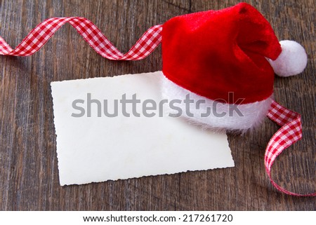 Santa Claus cap with vintage card and red-white checkered ribbon on dark wood, copy space