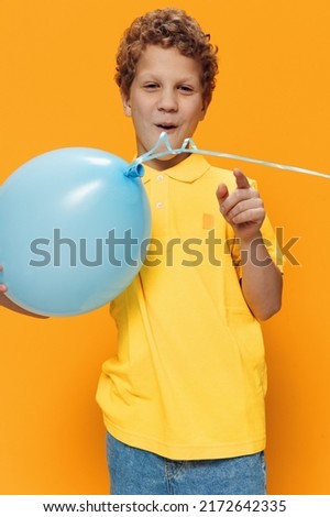 a cute, pleasant boy with curly hair stands on a yellow background holding a blue balloon in his hand