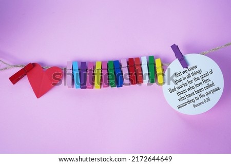 Bible verse quote - We know that in all things God works for the good of those who love him, who have been called according to his purpose. With colorful clips and red heart hanging on rope on pink.
