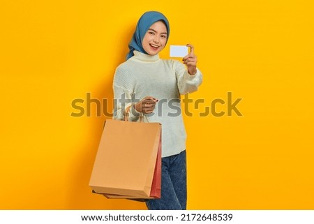 Cheerful beautiful Asian woman in white sweater holding shopping bags and showing credit card isolated over yellow background