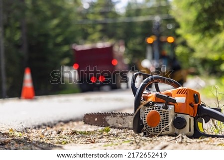 Close up selective focus view of a chainsaw on the verge of a main road. Blurry brake lights of an excavator and dumper truck are seen in background. Royalty-Free Stock Photo #2172652419