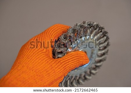 The hand in the orange glove holds the Disc Wire Brush. A tool for woodworking as well as stripping surfaces of corrosion, scale, or rust. First-person view. Gray background. Selective focus.