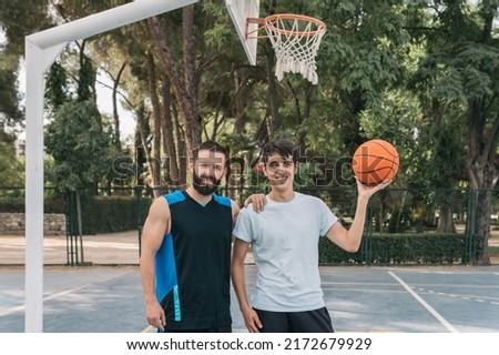 Smiling young boys under a basket with a ball on a blue court. A couple of sporty men, one of them holding an orange ball and the other leaning on his friend and rival. Concept of friendship and sport